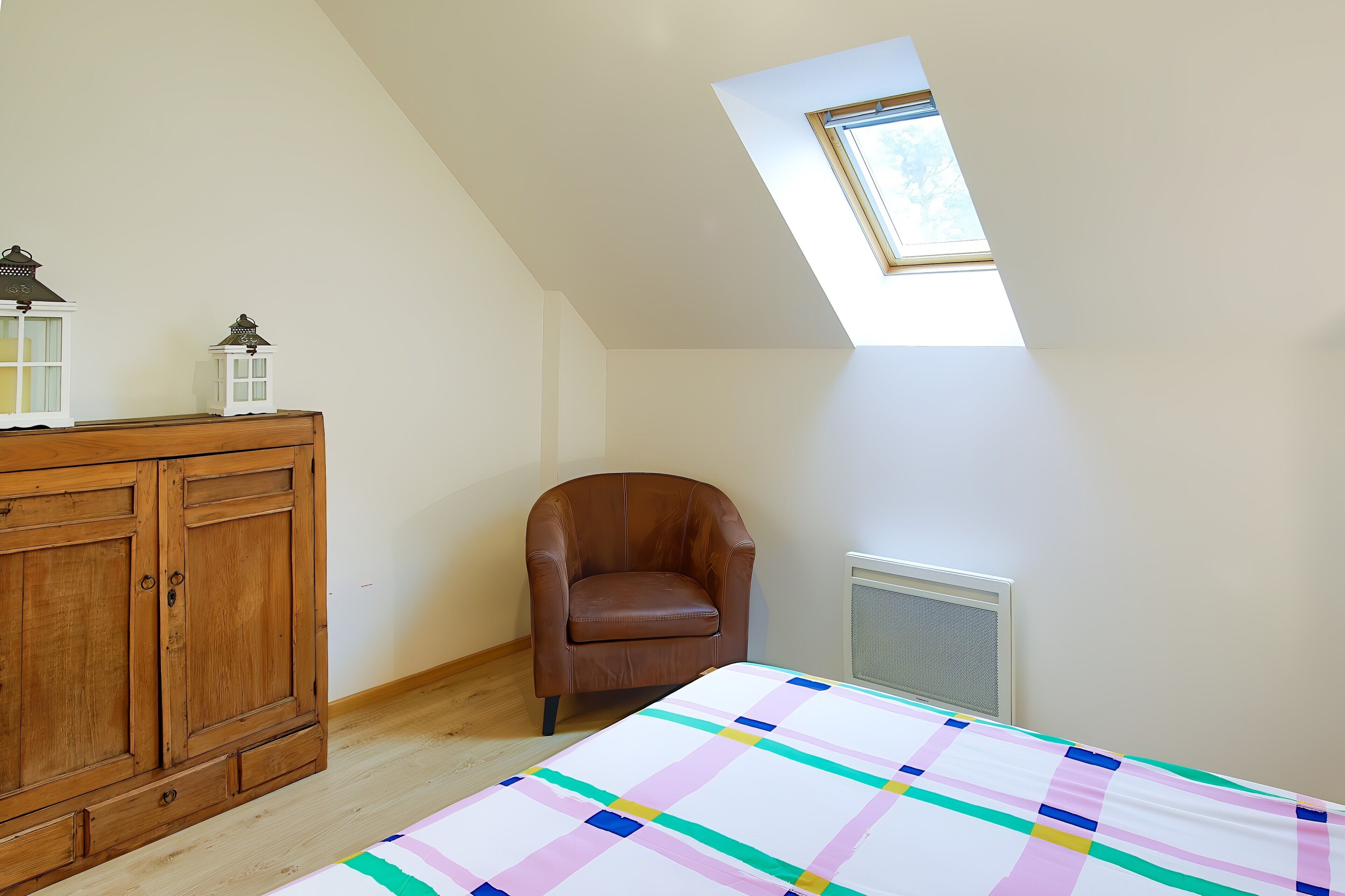 Een slaapkamer met een schuine kap, een bruine leren fauteuil, een houten dressoir en een kleurrijke quilt.