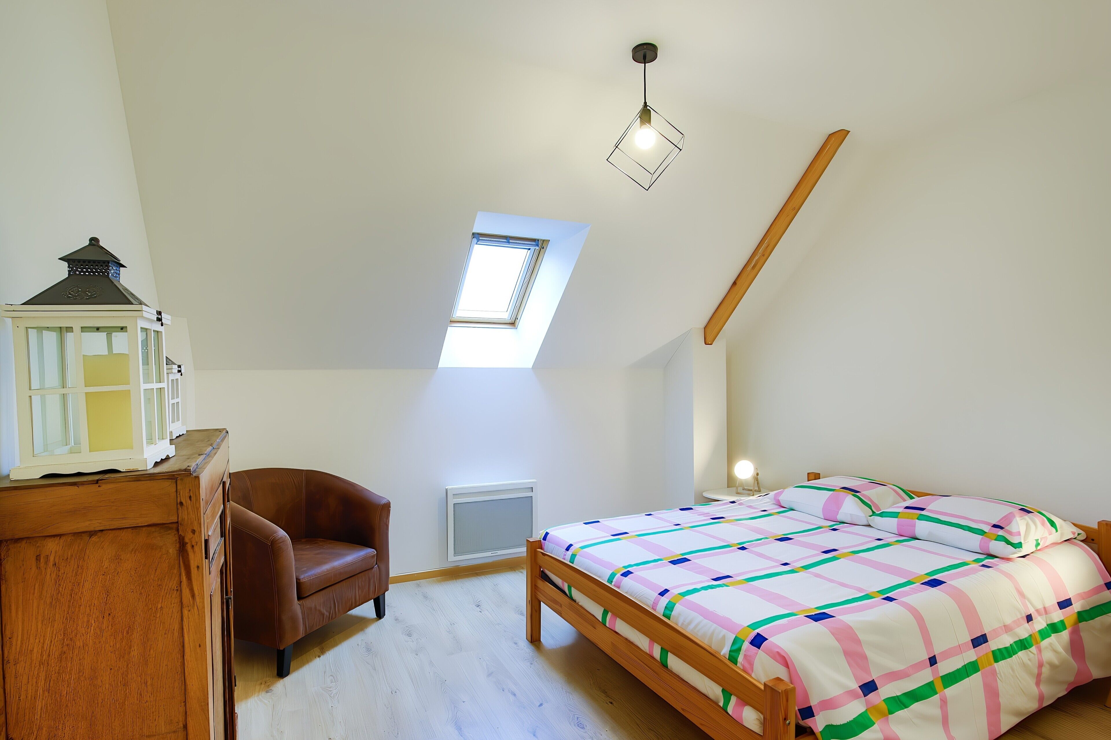 A bedroom with a sloped ceiling, a skylight, a bed with a colorful quilt, a wooden dresser, and a small white cabinet.