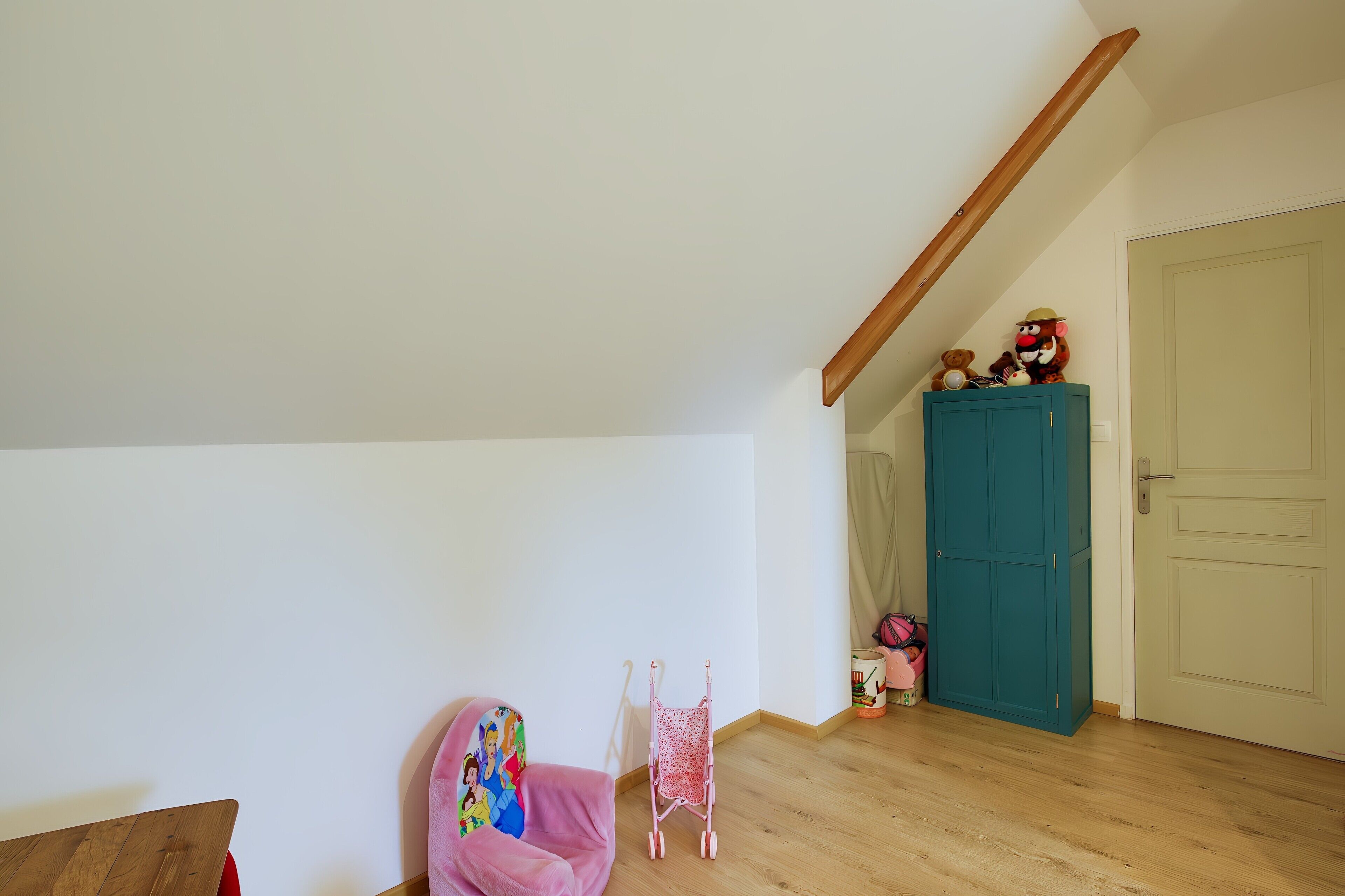 Una habitación con piso de madera, un armario azul verdoso, una silla rosa y un andador rojo pequeño.