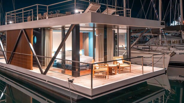 A modern floating structure with a glass-enclosed bedroom, wooden deck, and outdoor seating area. - Marina Suite (Loano)