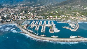 An aerial view of a coastal town with a marina, mountains in the background, and a rocky shoreline. - Marina Suite (Loano)