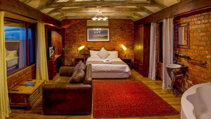 A bedroom with a bed, a red rug, a wooden bench, a bathtub, and a brick wall. - Madiba Bay Guesthouse (Gqeberha)