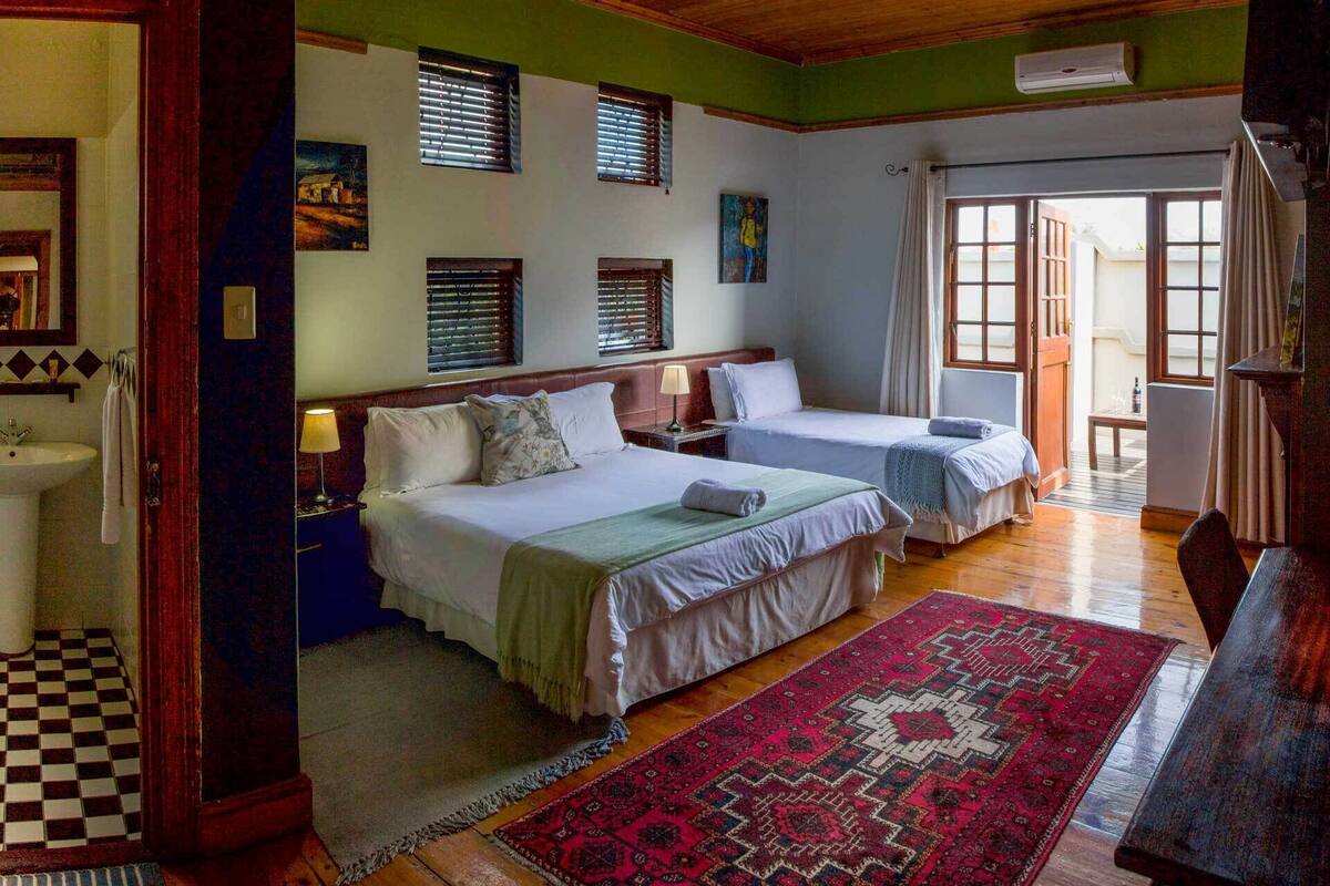 A room with two beds, a large window, a red patterned rug, and a wooden floor.