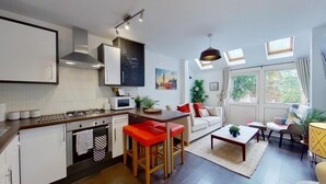 A compact kitchen with a gas stove, microwave, and a modern stove hood. There is a dining area with a sofa, red bar stools, and a coffee table. The room has large windows with curtains and a view of greenery outside. - Charming 2 Bed Apt with Netflix & Wi-Fi (London)