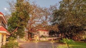 A paved courtyard with a roundabout, surrounded by lush greenery and trees, leading to a building with a red roof. - Staymaster Terra Tango 4BR Villa (Calangute)