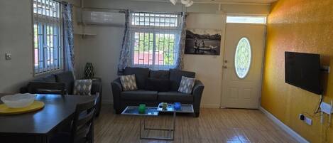 A living room with a sofa, coffee table, dining table, and chairs. There is a ceiling fan, a door with a glass window, and a wall-mounted TV.