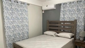 A bedroom with a wooden bed, patterned curtains, an air conditioning unit, and a nightstand with a lamp. - Travellers Villa (Roseau)