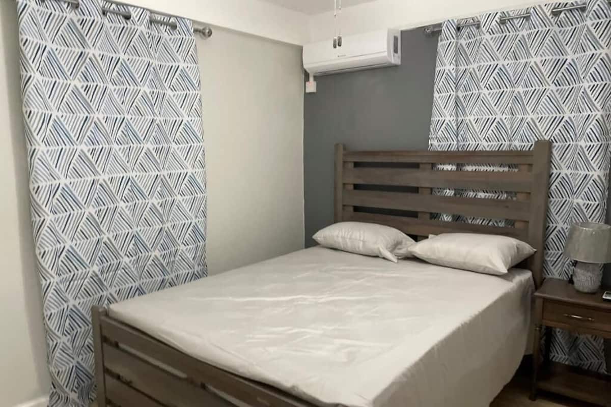 A bedroom with a wooden bed, patterned curtains, an air conditioning unit, and a nightstand with a lamp.