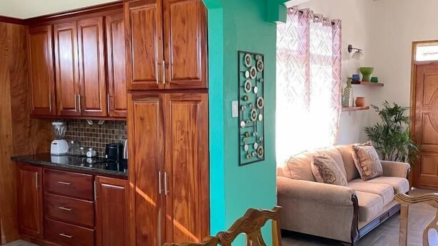 A kitchen with wooden cabinets, a dining table with chairs, a sofa, and a decorative wall piece. - Travellers Villa (Roseau)
