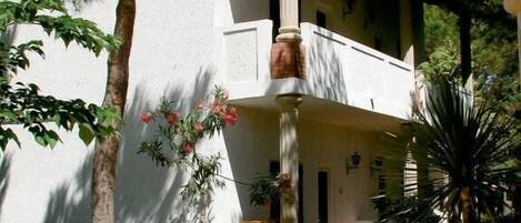 A white building with a balcony, surrounded by trees and plants.