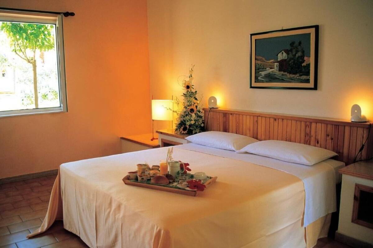 A hotel room with a bed, a tray of breakfast items, a nightstand, a lamp, a wall-mounted painting, and a window with a view of trees.