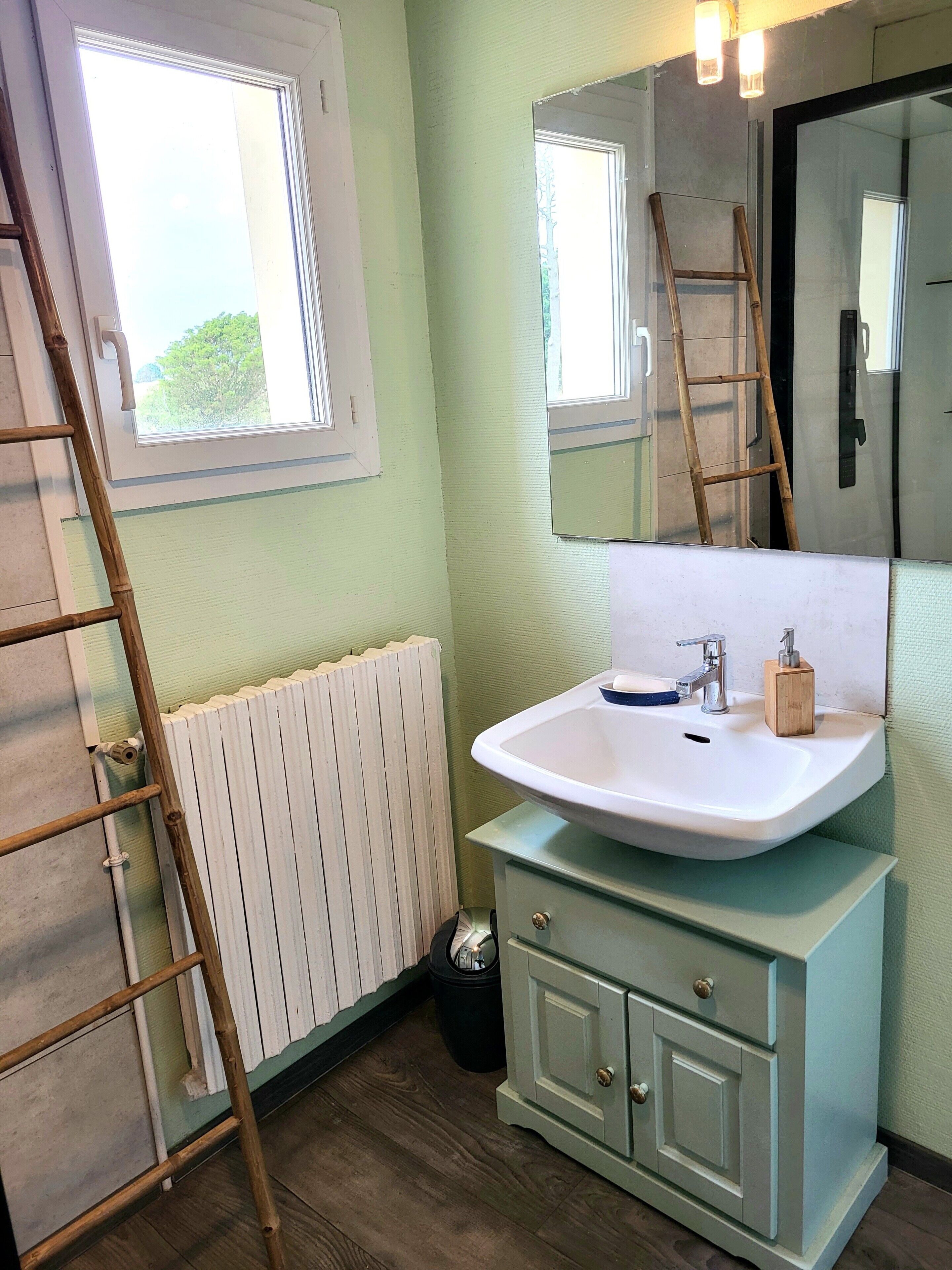 Een badkamer met een witte radiator, een wastafel met een spiegel erboven, een houten ladder en een raam.