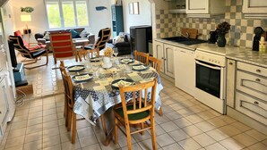 A dining area with a table set for four, a kitchen with white cabinets, and a living room with a sofa and chairs. - Seaside, Les Fleurs Holiday Home with Shared Terrace, Private Garden and Wi-Fi (Créances)