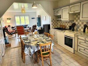 Un comedor con una mesa dispuesta para cuatro, una cocina con armarios blancos y una sala de estar con sofá y sillones.