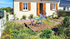 A single-story house with a front porch, a small garden with various plants and flowers, and a wooden deck. - Seaside, Les Fleurs Holiday Home with Shared Terrace, Private Garden and Wi-Fi (Créances)