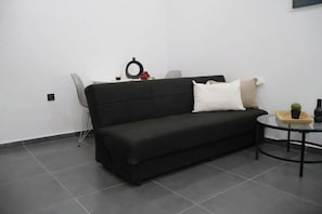 A black sofa with white and beige pillows, a small round table with a plant, and a chair in a minimalistic room with white walls and grey flooring. - KAPETAN MATAPA 55, PIREAUS, 185 44 (Pireas)