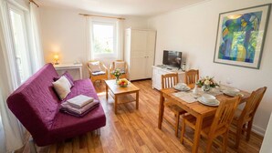 Una sala de estar acogedora con un sofá morado, una mesa de comedor de madera y sillas, un televisor y un cuadro en la pared.