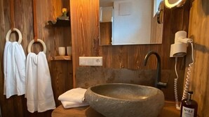 Een badkamer met een stenen wastafel, houten wandpanelen, een spiegel met ronde verlichting en twee handdoeken aan een handdoekhouder.