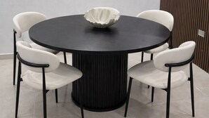 Uma mesa de jantar redonda com um arranjo de uma tigela branca no centro, rodeada por quatro cadeiras brancas com pernas pretas, colocada diante de uma parede de cor clara com uma pintura abstrata texturizada acima.
