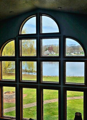 Une pièce dotée de grandes fenêtres en arc qui offrent une vue sur un lac, une zone herbeuse et des maisons situées au loin.