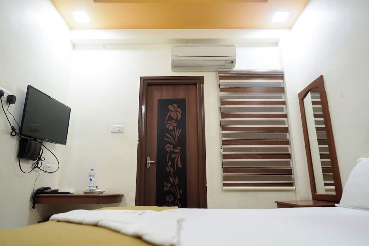A hotel room with a bed, a TV mounted on the wall, an air conditioning unit, a door with a floral design, and a window with blinds.