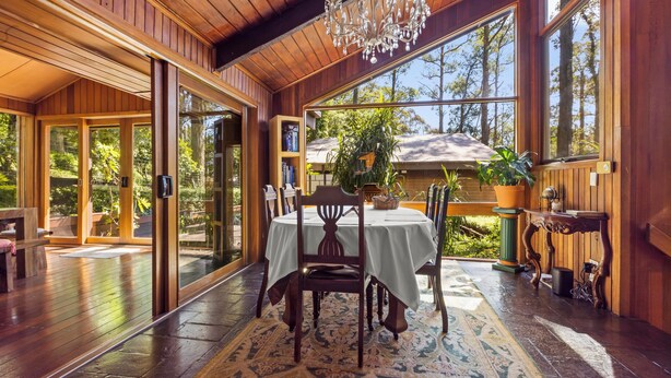 A wooden interior with a dining table, chairs, a chandelier, and large windows.