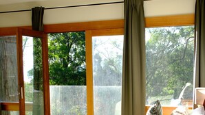 A bedroom with a bed, a bedside table with a lamp, a window with curtains, and a view of greenery outside.