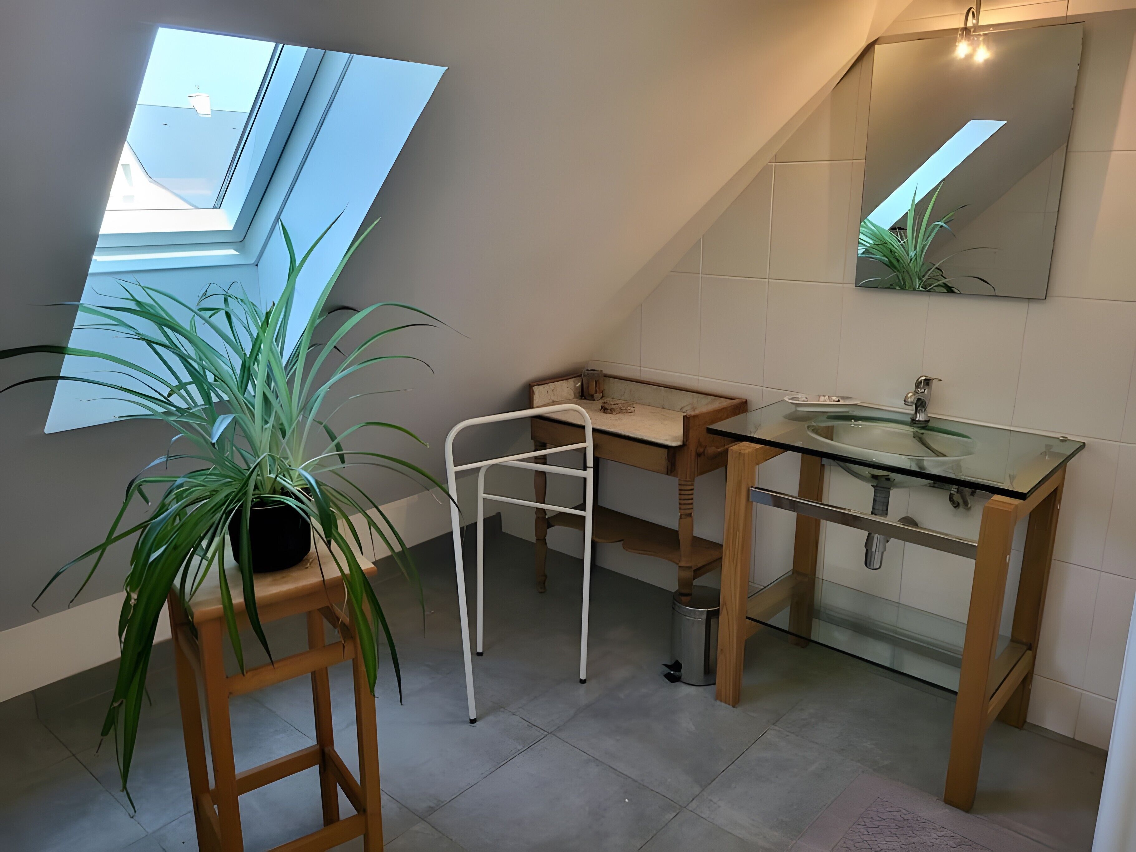 Um banheiro moderno com claraboia, pia de vidro, toucador de madeira e planta em vaso.
