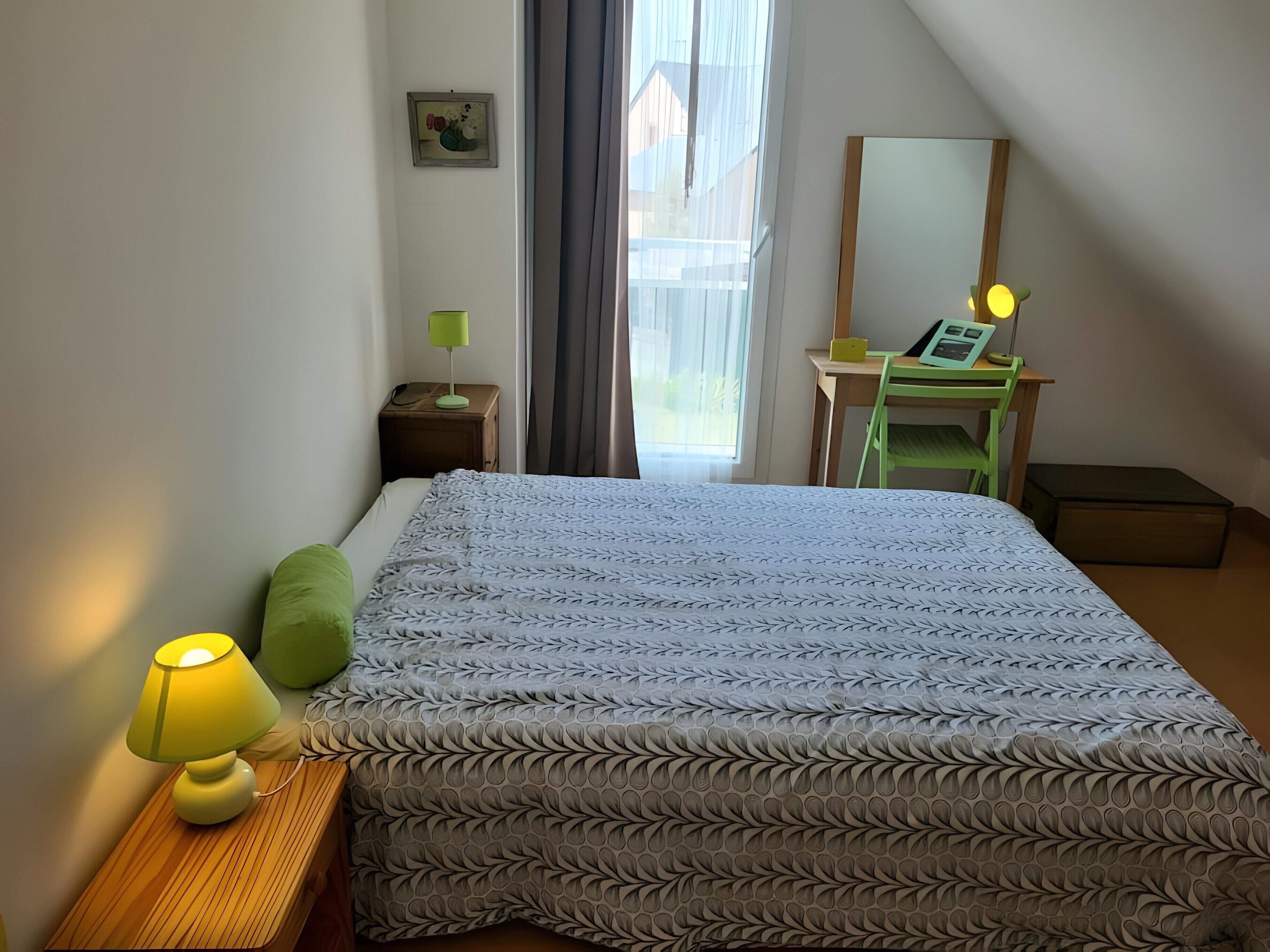 Un dormitorio con cama, lámparas de noche, escritorio, silla, espejo y ventana con cortinas.