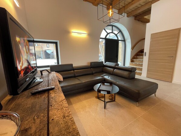 Un salon moderne avec un canapé d’angle, un téléviseur à écran plat, une table basse en bois et une grande fenêtre offrant une belle vue.