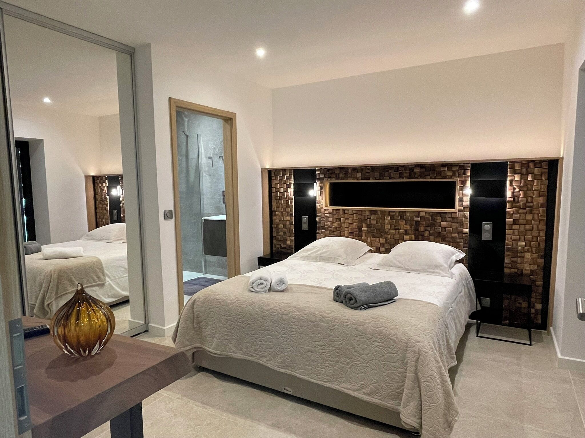 A modern hotel room with a large bed, a bedside table, a mirror, and a wall-mounted TV.
