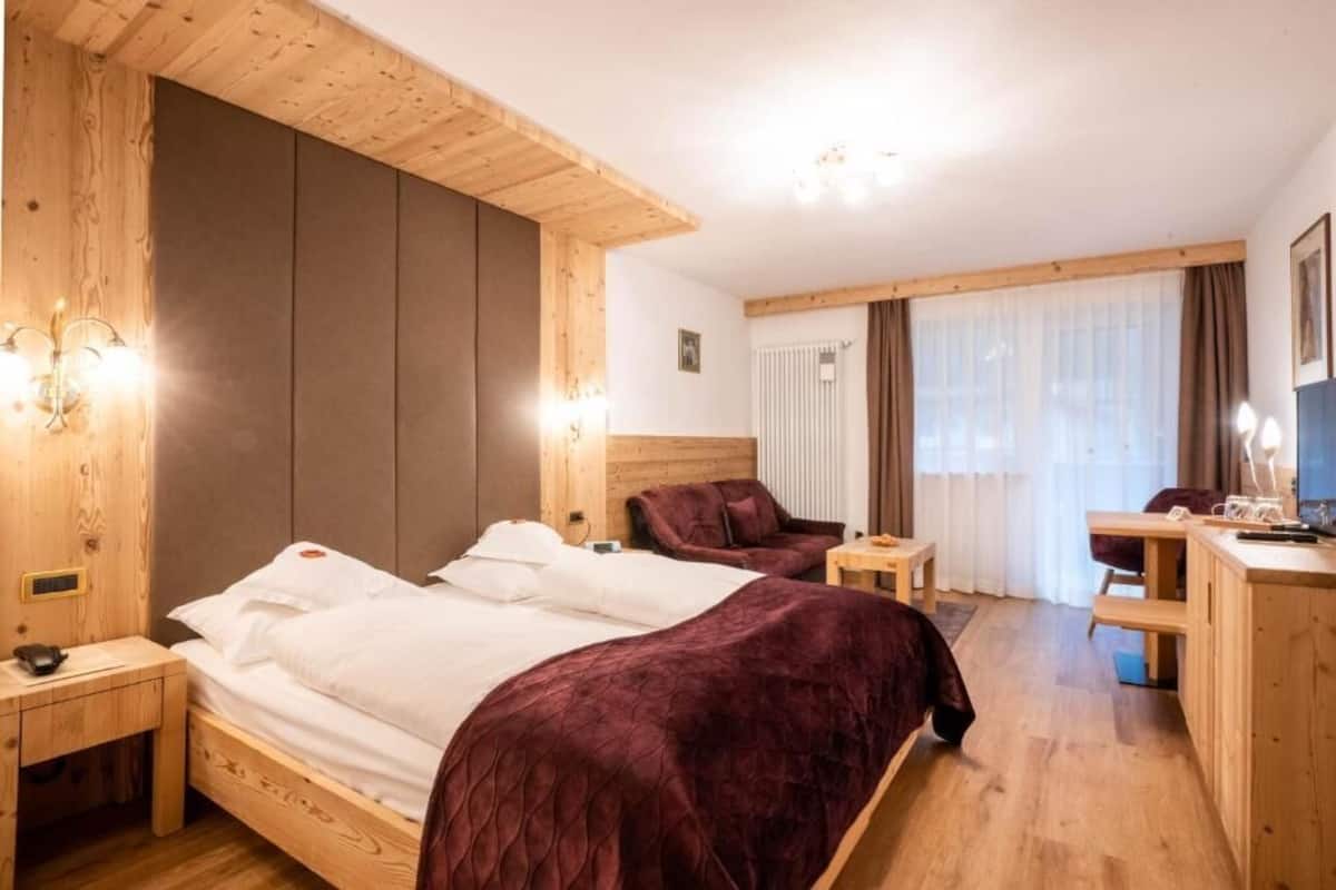 Habitación de hotel con una cama de madera, un escritorio, una silla y un ventanal con cortinas.