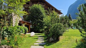 Un chemin de pierres mène à un bâtiment en bois doté de balcons, le tout étant entouré d’une végétation luxuriante et ayant une chaîne de montagnes en toile de fond.