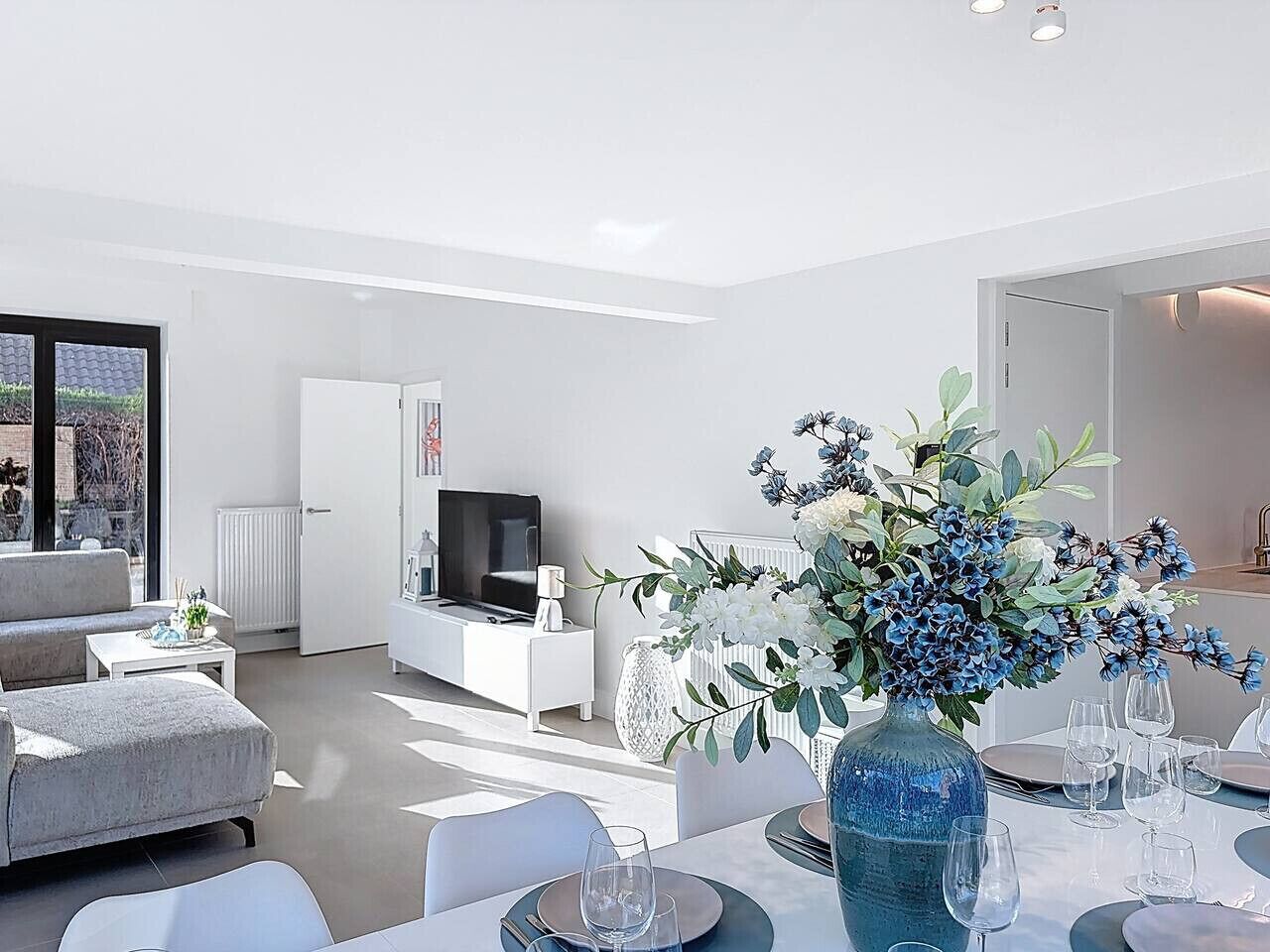 A modern living room with a sofa, TV, dining table set, and a vase of flowers.