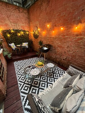 A cozy patio with a brick wall, string lights, a patterned rug, a dining table with chairs, and a sofa with cushions. - Chelsea 2BR Private Courtyard Earl s Court Tube (London)