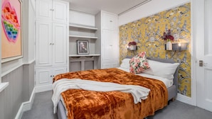 A bedroom with a large bed, white built-in wardrobe, a decorative wall, and a small shelf with a vase. - Chelsea 2BR Private Courtyard Earl s Court Tube (London)