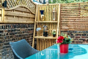 A glass table with a wicker chair, a wooden shelf with decorative items, and a brick wall. - Kensington Chelsea 1BR Patio Apartment Tube (London)