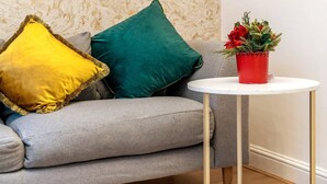 Un sofá gris con dos almohadas, una maceta roja con flores y una mesa blanca con patas doradas.