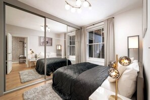 A modern bedroom with two beds, a large mirror, a bedside lamp, and a view of the adjacent room. - Kensington Chelsea 1BR Patio Apartment Tube (London)