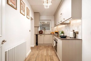 A narrow kitchen with white cabinets, a wooden floor, and a window with a wreath. - Kensington Chelsea 1BR Patio Apartment Tube (London)
