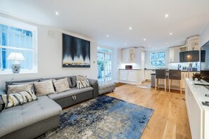 A modern living room with a grey sofa, a large abstract painting, and a kitchen area with white cabinets and a dining table. - Kensington Chelsea 2BR Garden Apartment Tube (London)