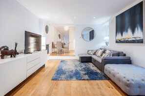 A modern living room with a sofa, a television, and a dining area in the background. - Kensington Chelsea 2BR Garden Apartment Tube (London)