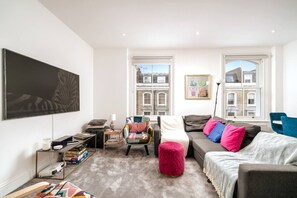 A living room with a sofa, ottoman, armchair, and a large painting on the wall. - Chelsea 5BR Family Home Walk to Earl s Court Tube (London)