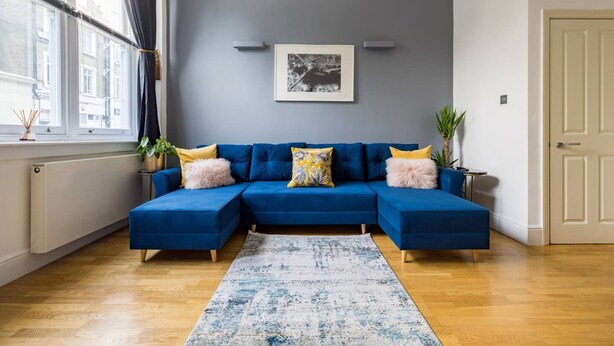 Ein modernes Wohnzimmer mit einem blauen Modulsessel, einem gerahmten Bild an der Wand und einem Fenster mit Vorhängen.