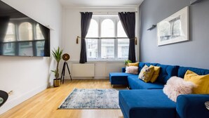 Ein Wohnzimmer mit einem blauen Sofa, einem großen Fenster mit Vorhängen, einem Stativ und einer Pflanze.
