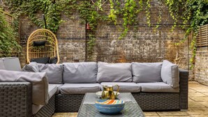 Eine Rattan-Couch, ein Rattan-Sessel, eine gemusterte Schale und eine goldene Kanne auf einer gefliesten Terrasse.