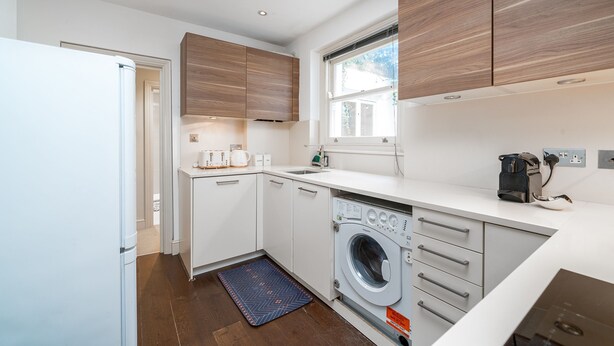 A compact kitchen with a washing machine, a coffee maker, and wooden cabinets. - Chelsea 2BR Garden Patio Apartment Tube (London)