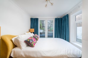 A bedroom with a bed, a yellow armchair, a lamp, and a view through a glass door. - Maida Vale 2BR Stylish Garden Apt Walk Tube (London)