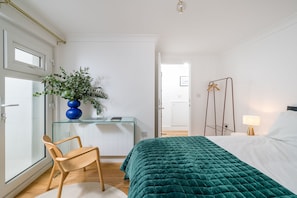 A bedroom with a bed, a chair, a desk with a plant, a hanging light, and a door. - Maida Vale 2BR Stylish Garden Apt Walk Tube (London)