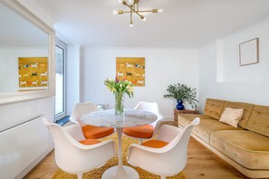 Una sala de estar moderna con una mesa redonda de mármol, sillas blancas con cojines naranjas, un sofá beige y un ventanal grande.